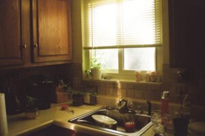 A pile of dishes and pans sits in a kitchen sink in the dim sunlight, waiting to be washed.